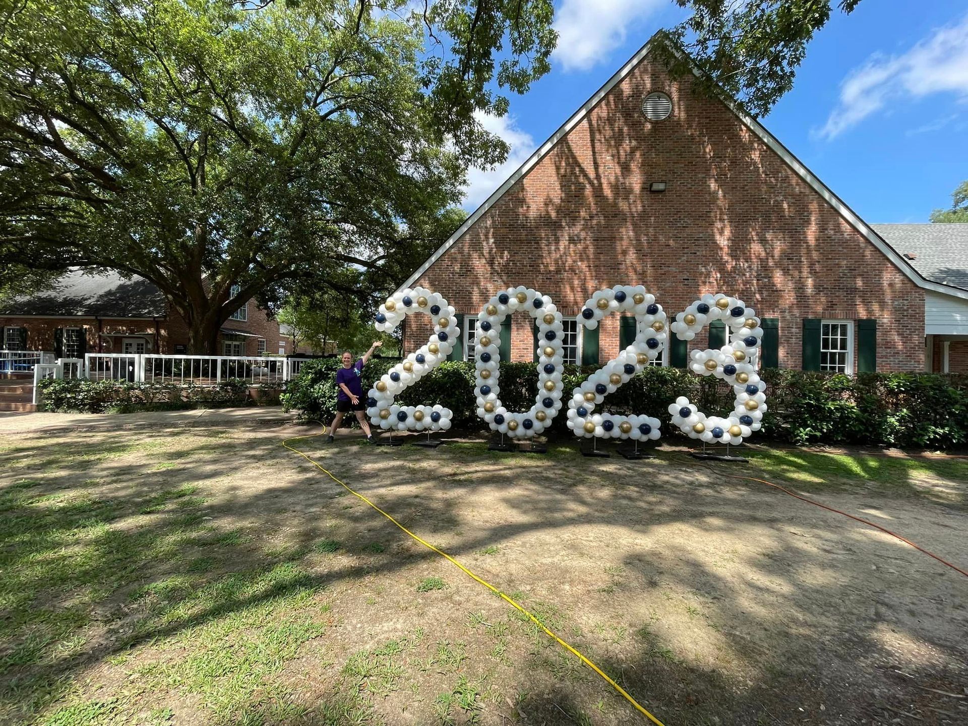 A man is standing in front of a brick house with balloons in the shape of the year 2023.