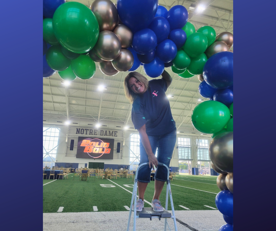 A woman is standing on a ladder decorating balloons in a stadium.