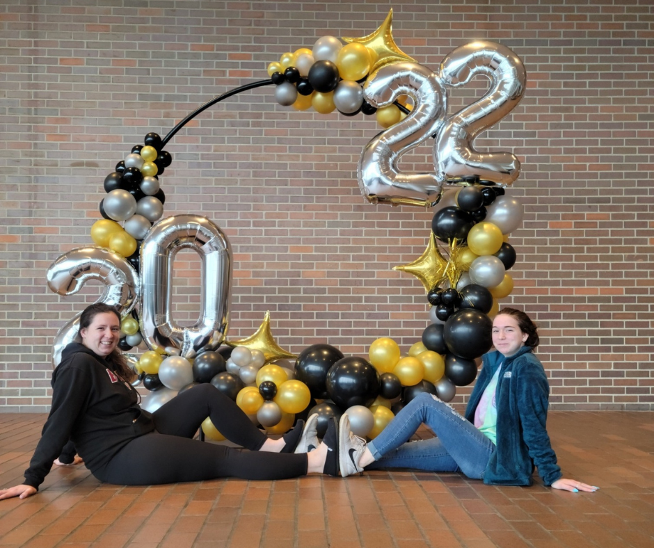 Two women are sitting on the floor in front of a balloon infinity hoop photo frame