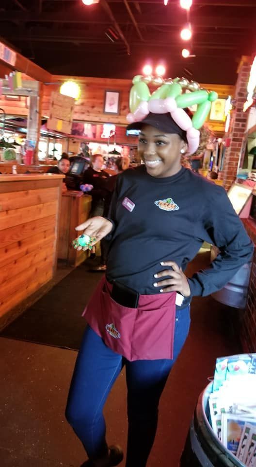 A woman wearing a frog balloon hat is standing in a restaurant.