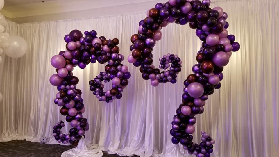A large arch made of purple and black balloons is sitting in front of a white curtain.