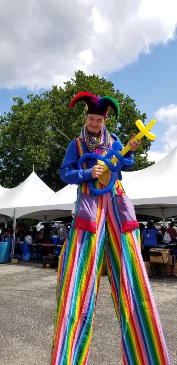 A clown is standing on stilts holding balloons and a guitar.