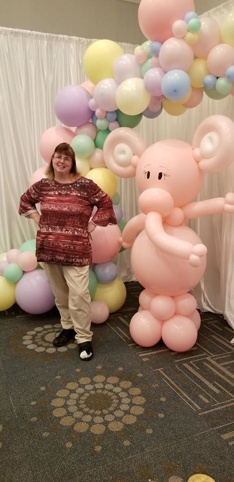A woman is standing next to a balloon sculpture of an elephant for a baby shower.