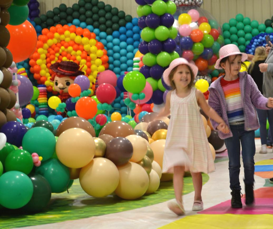 Two little girls are walking in front of a wall of balloons. Candy Land