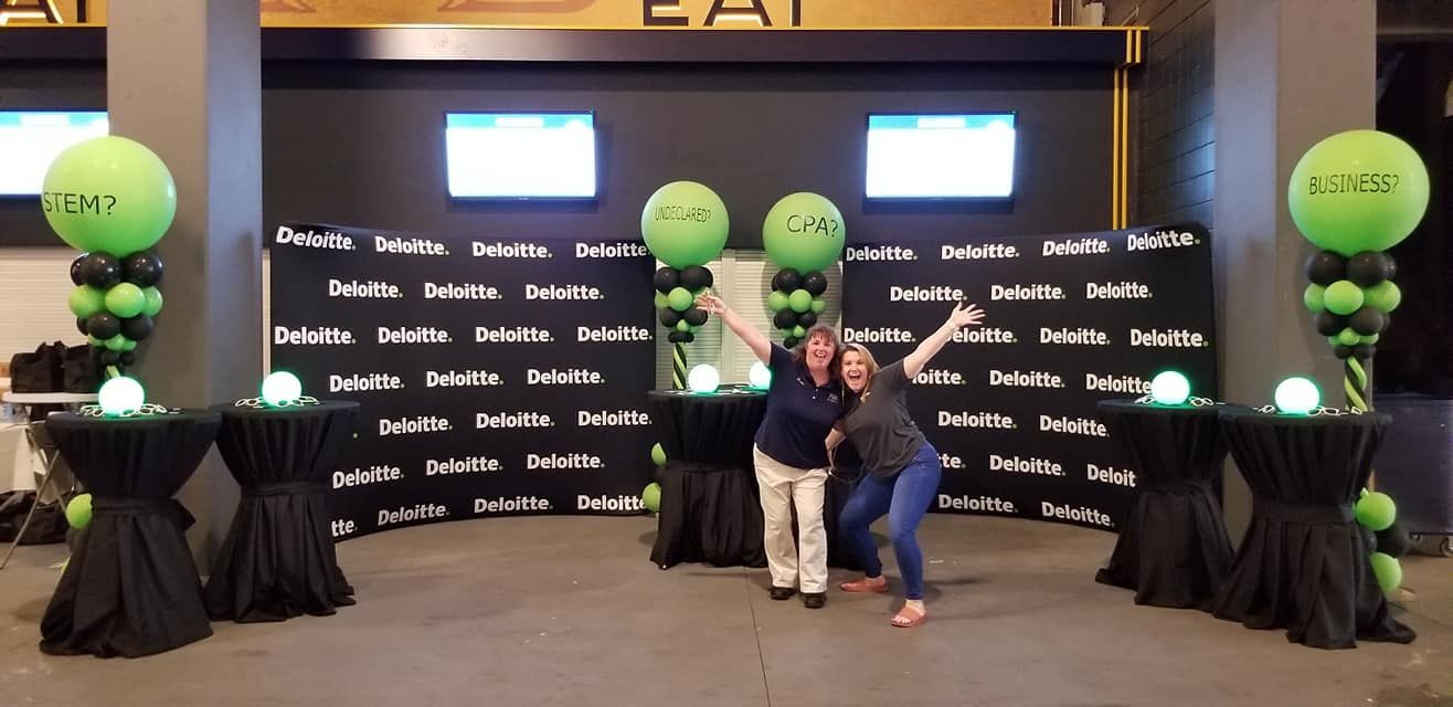 Two people are standing in front of a wall with green balloons.