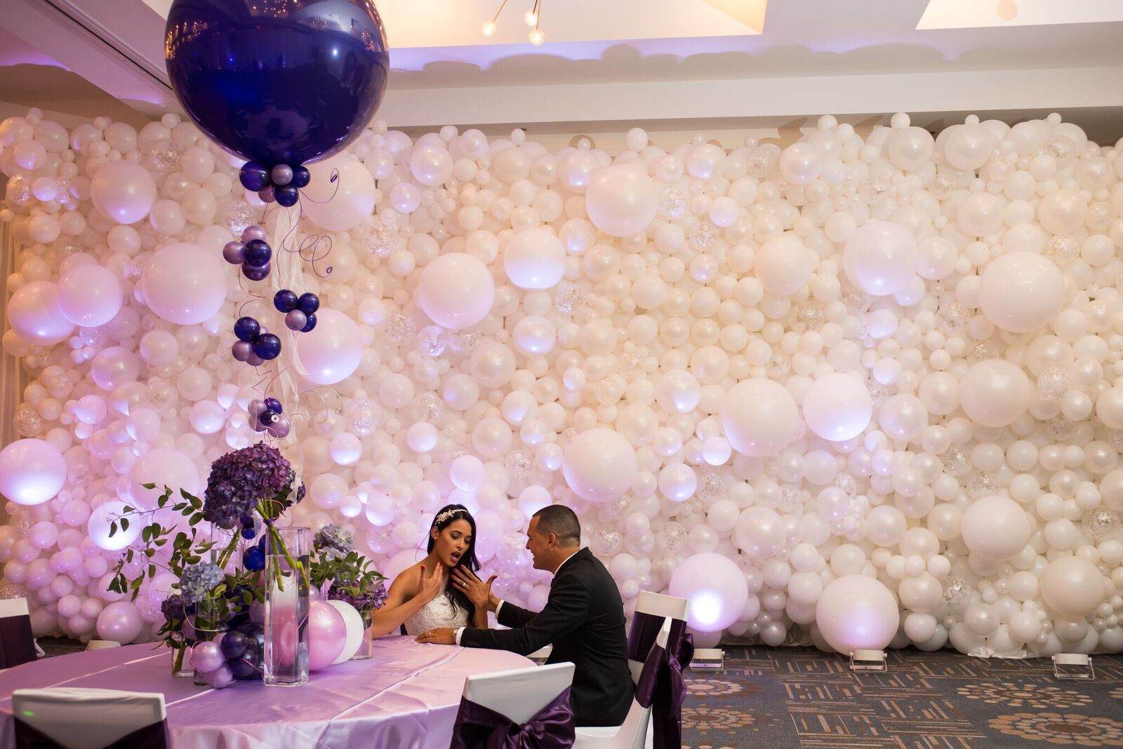 A bride and groom are sitting at a table in front of a wall of balloons.
