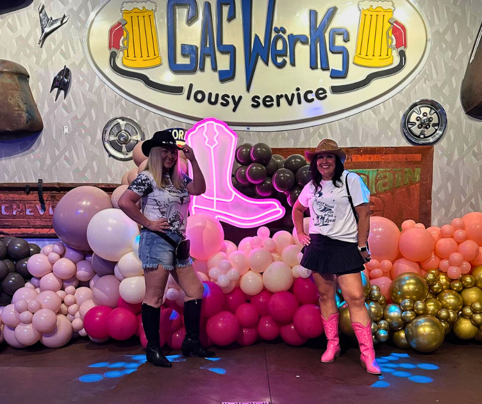 Two women posing in front of a balloon arch and neon boot. Gaswerks sign in the background.