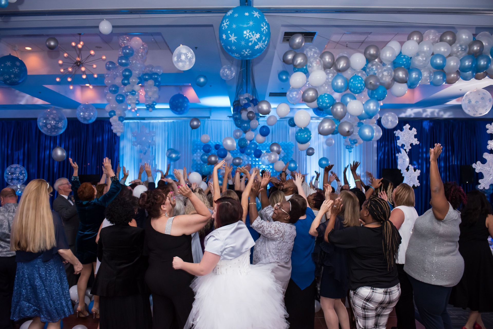 A large group of people are dancing in a room with balloons hanging from the ceiling.