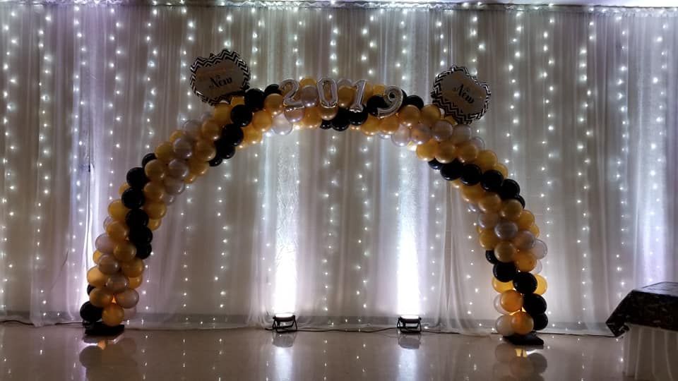 A black and gold balloon arch is sitting in front of a white curtain.