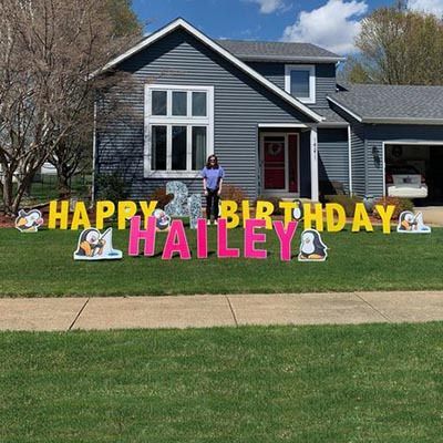 A woman is standing in front of a house with a happy birthday sign.