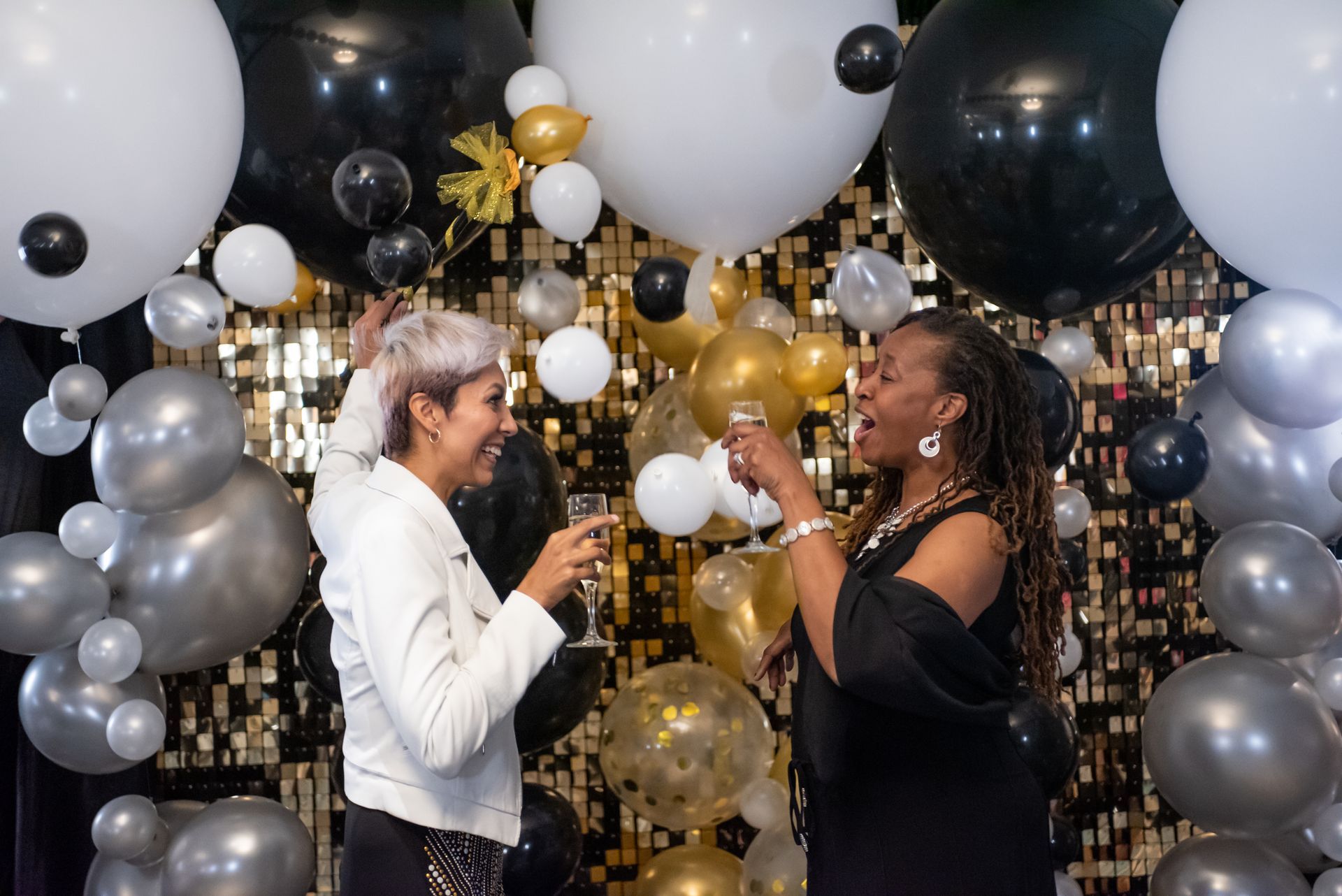 Two women are toasting with champagne in front of a wall of balloons.