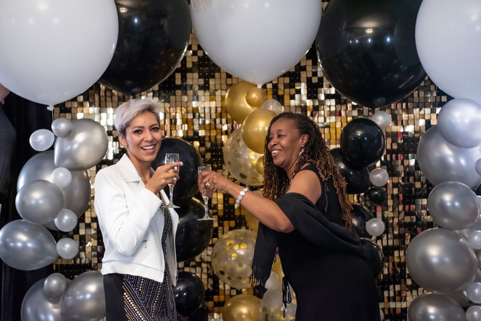 Two women are toasting with champagne in front of a wall of balloons.