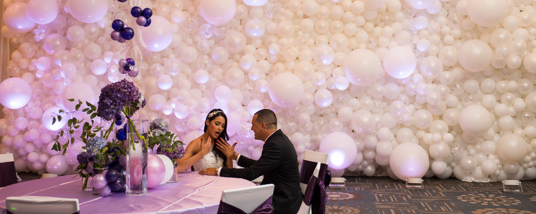A bride and groom are sitting at a table in front of a wall of balloons.