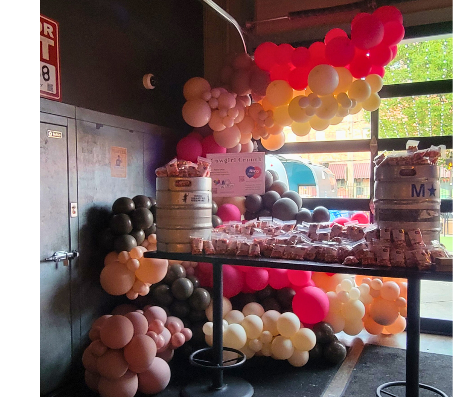 Party scene with a table of food, beer kegs, and balloon arch in various colors.