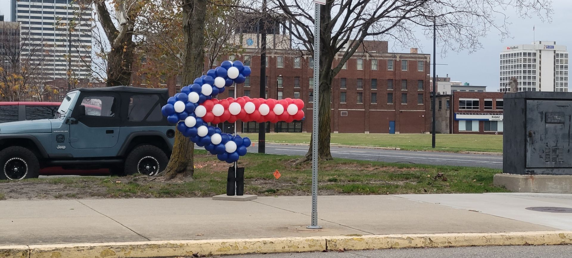 A balloon arrow in red, white, and blue points left, near a blue Jeep in front of a brick building.