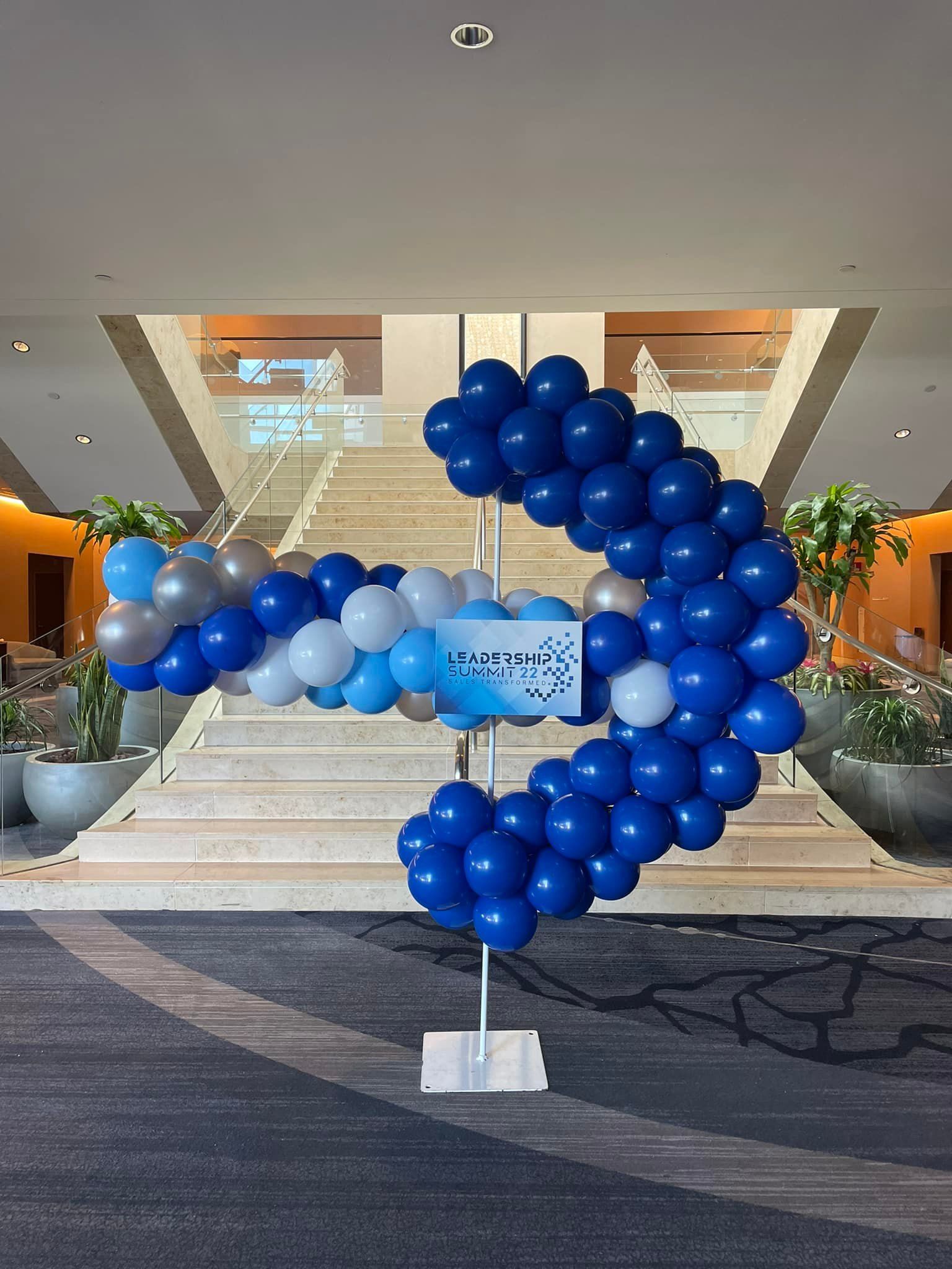 Blue, silver, and white balloon archway over a stand in a grand hall with stairs.