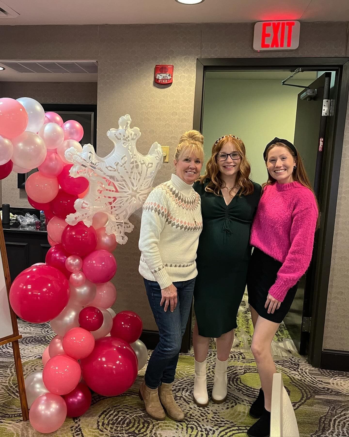 Three women are posing for a picture in front of a red exit sign.