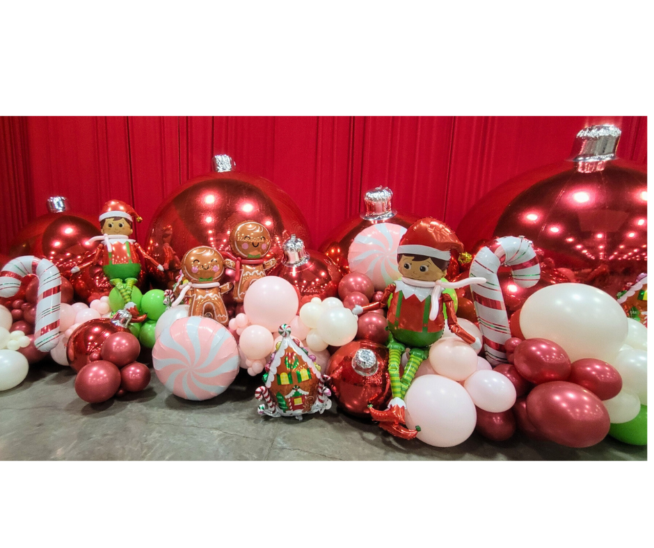Christmas balloon display with ornaments, gingerbread figures, and elves on a red backdrop.