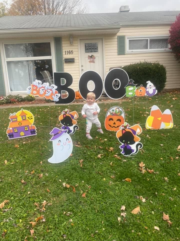 A baby is standing in front of a house decorated for halloween. Boo themed yard card.