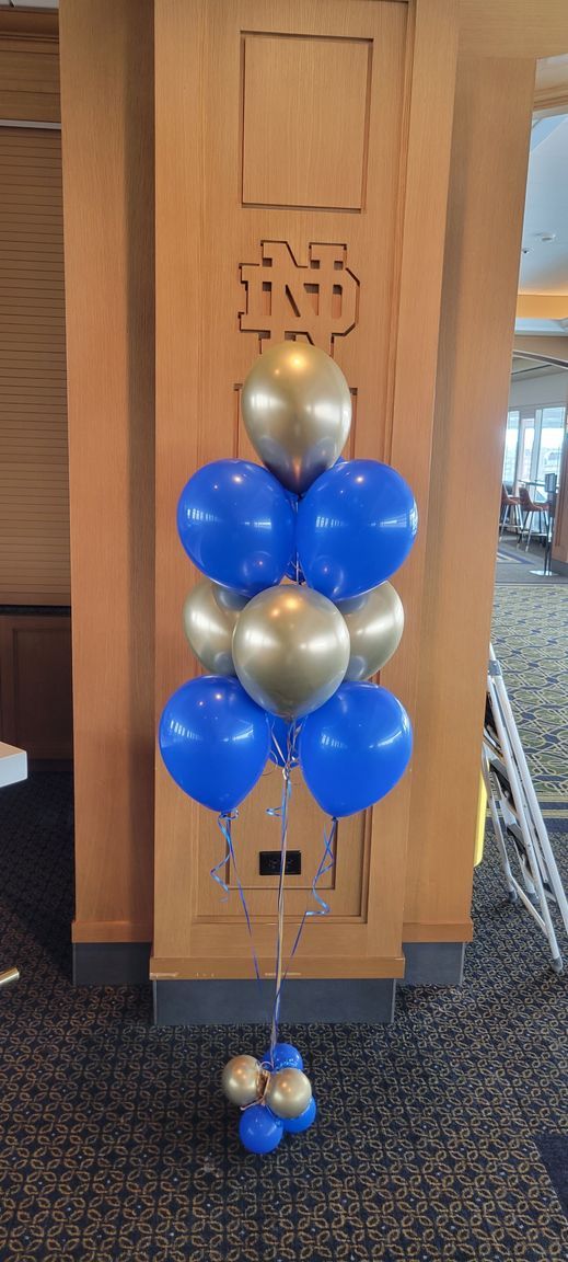 A balloon arrangement in blue and gold. The balloons are in front of a wooden panel with a logo.