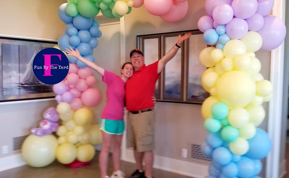 A man and a woman are standing in front of a rainbow of balloons.