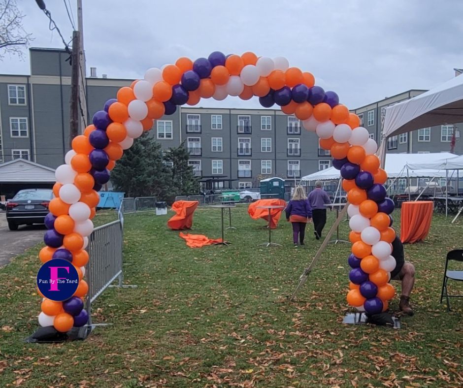 Balloon arch in orange, purple, and white. People walk through. Outdoor event.