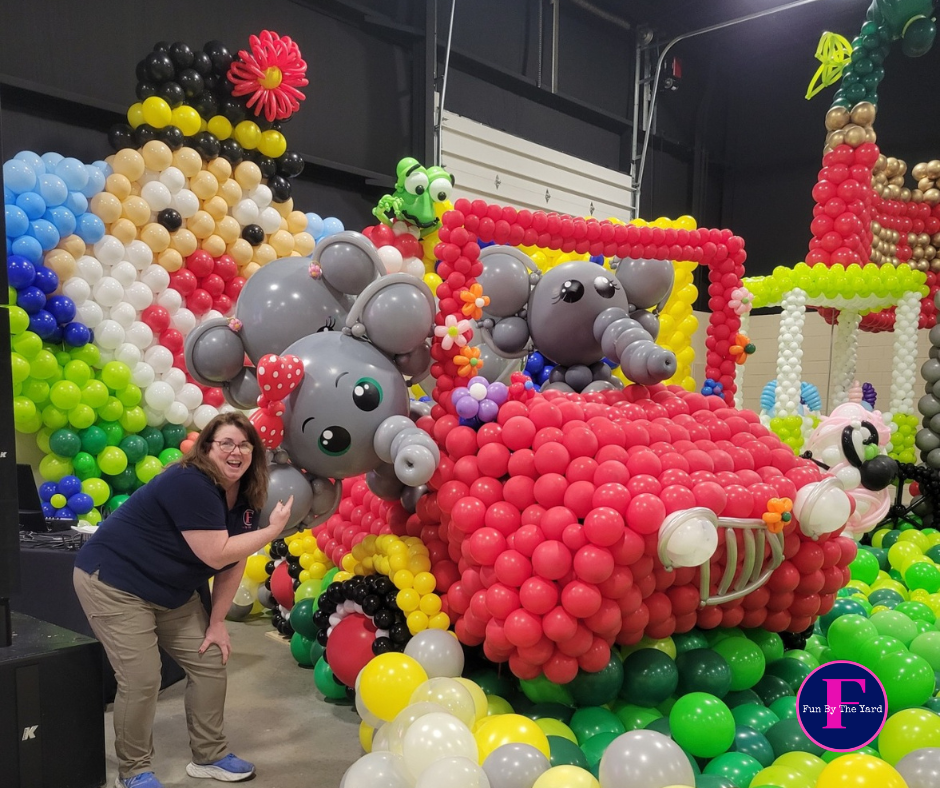A woman is standing in front of a car of elephants made out of balloons. Fun By The yard