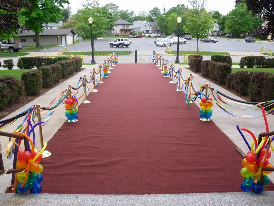 Red carpet leading to a building, lined with rope and balloon decorations, outdoor setting.