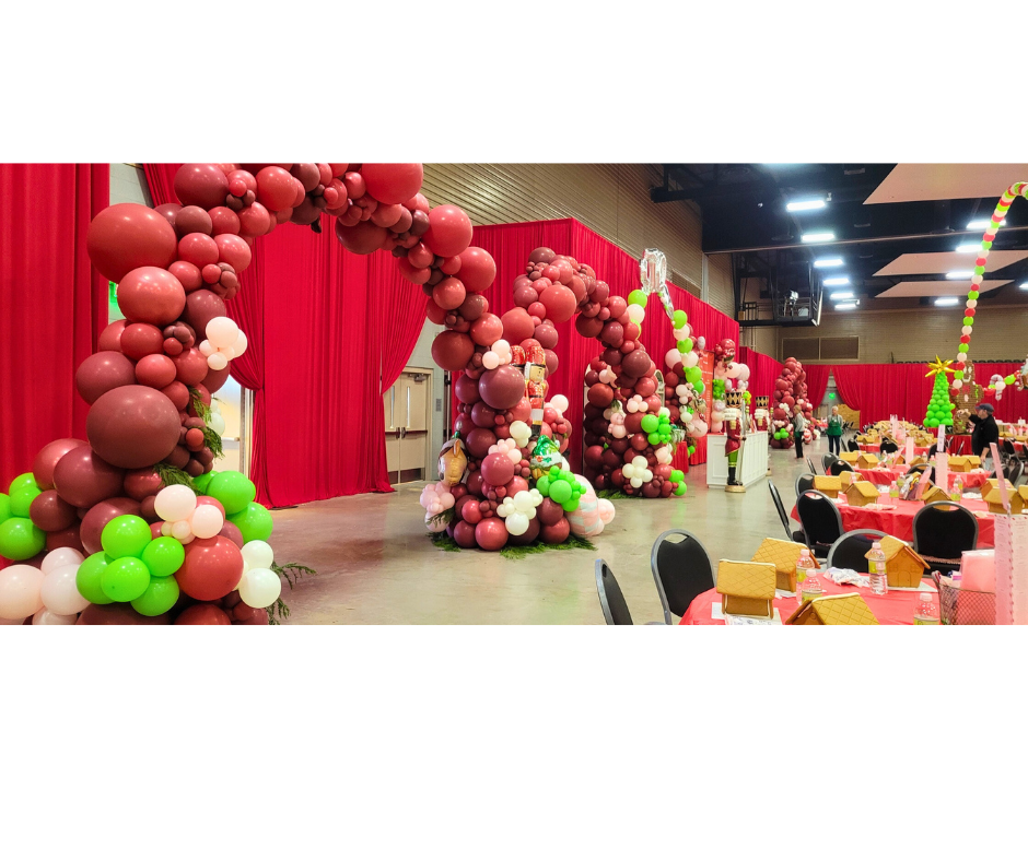 Red and green balloon arches decorate a holiday event space with tables and red curtains.