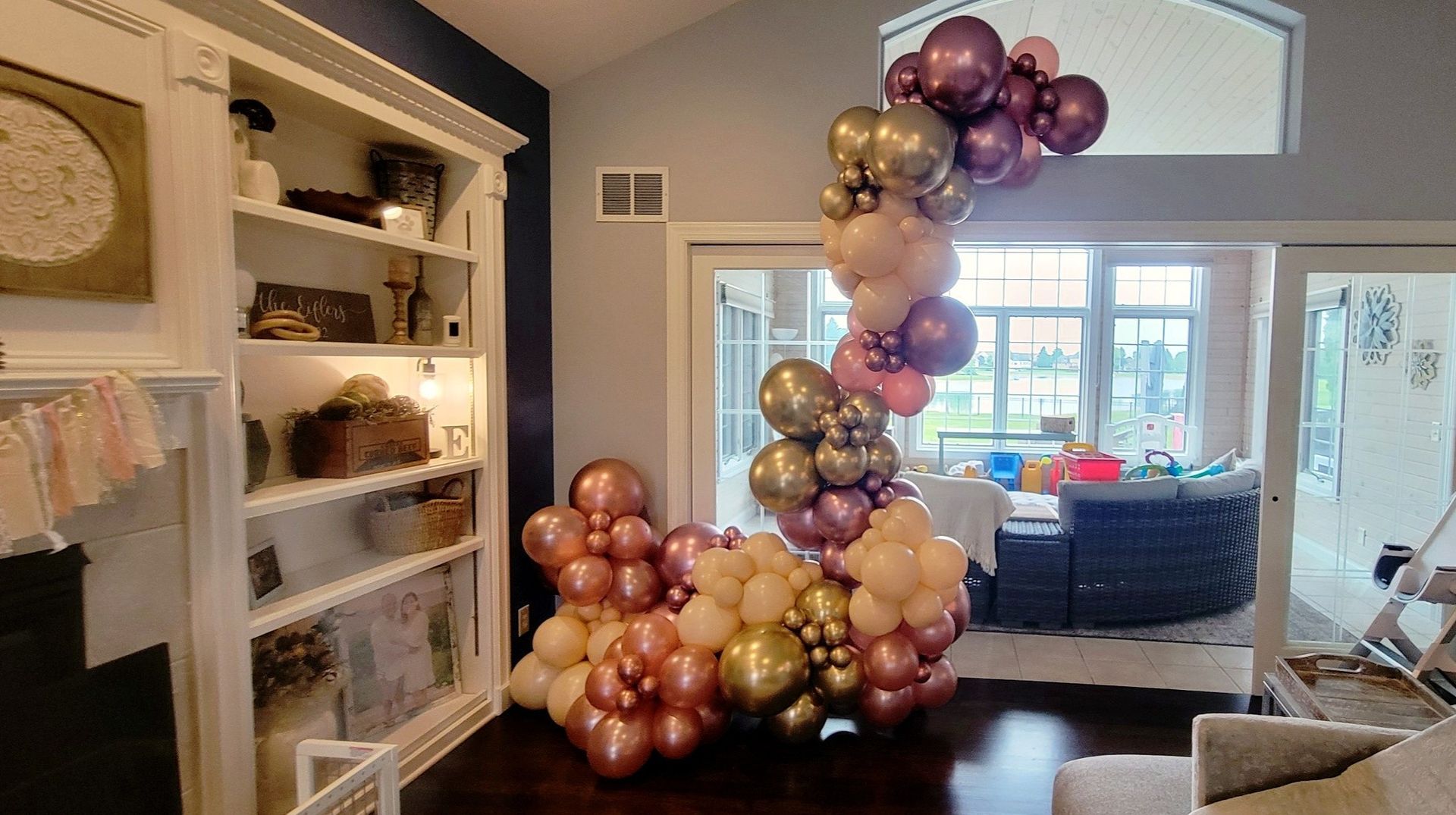 Balloon arch with gold, rose, and purple balloons in a living room.