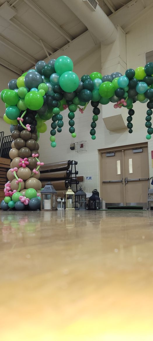 A decorative balloon arch in a school gymnasium; the arch is green and brown and shaped like a tree.