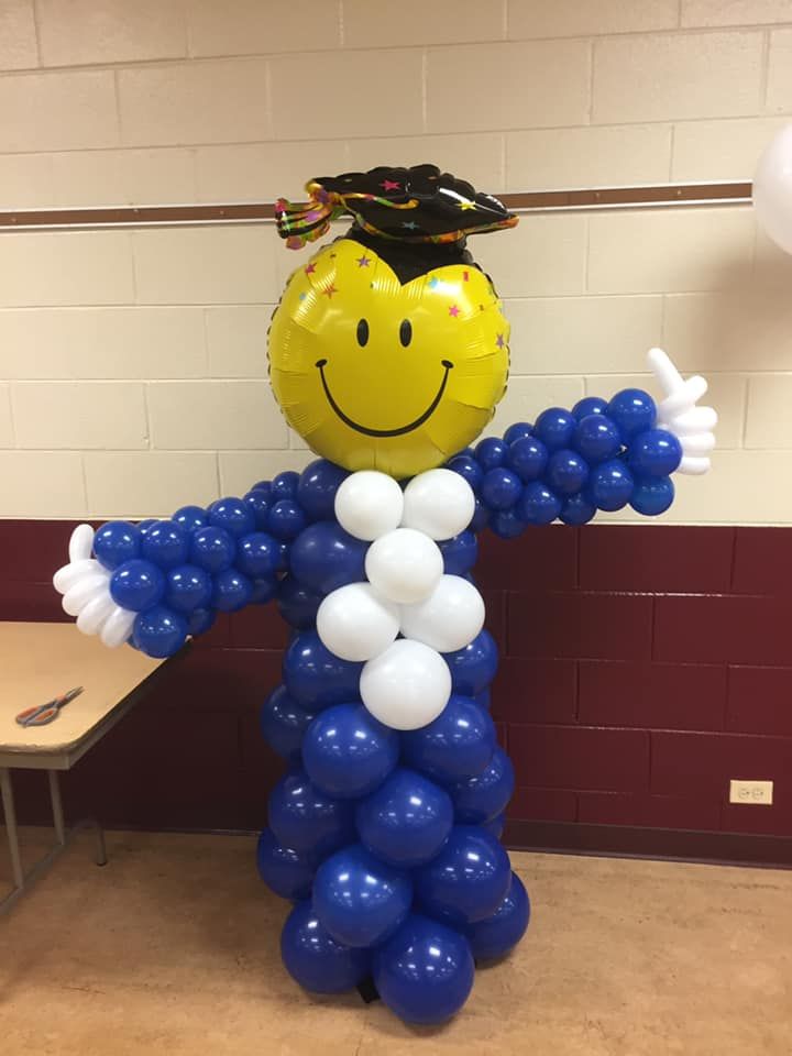 A yellow balloon with a smiley face on it is wearing a graduation cap and gown made of blue and white balloons