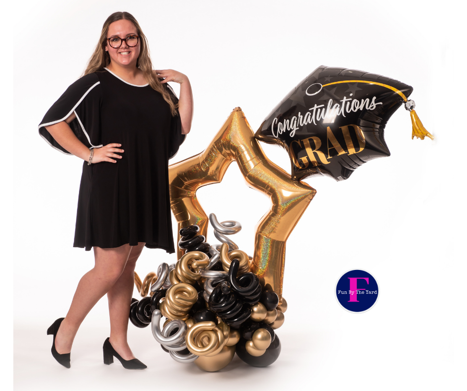 A woman in a black dress is standing next to a graduation cap balloon.