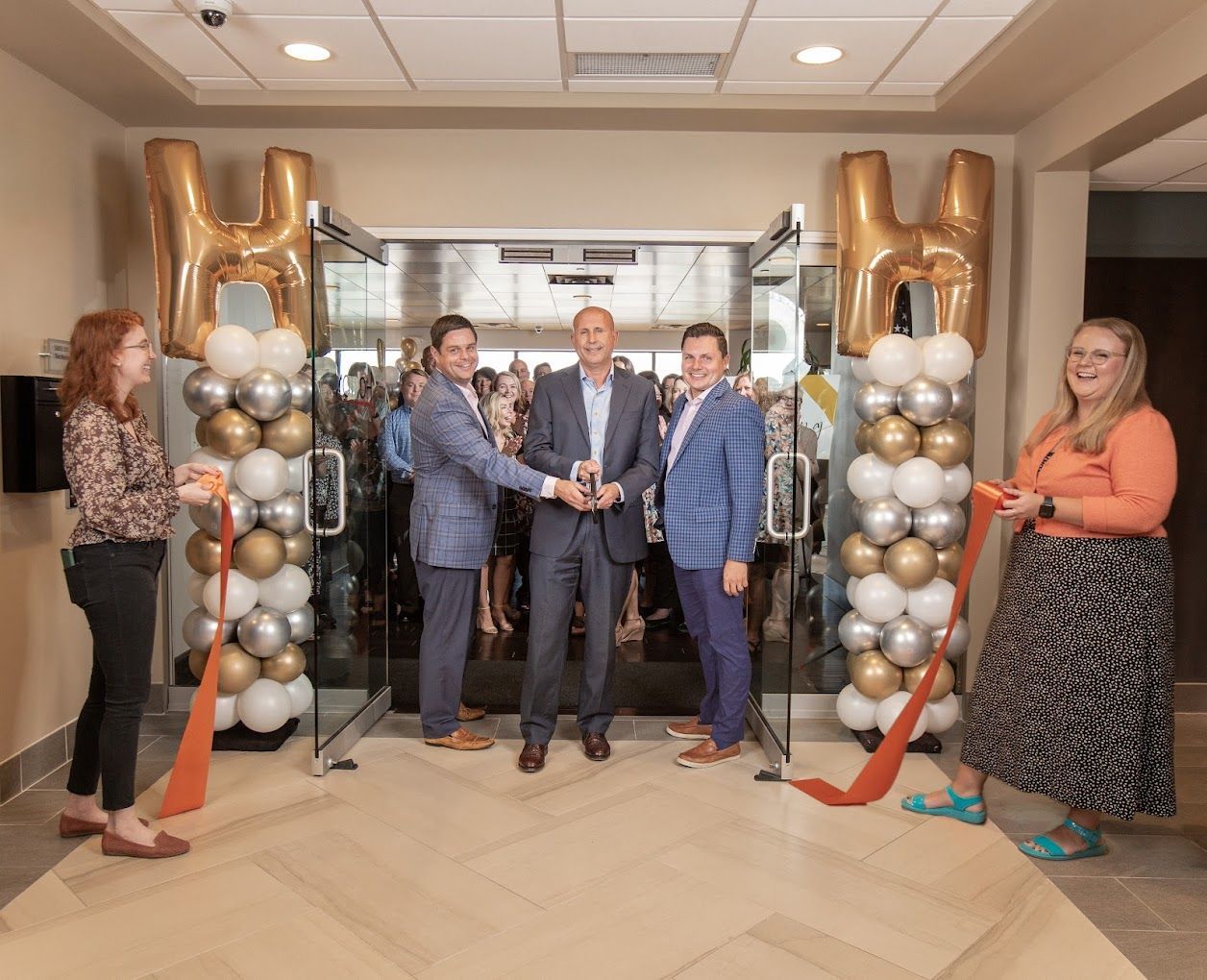 People cut a ribbon at a doorway, flanked by balloon columns, for a grand opening.
