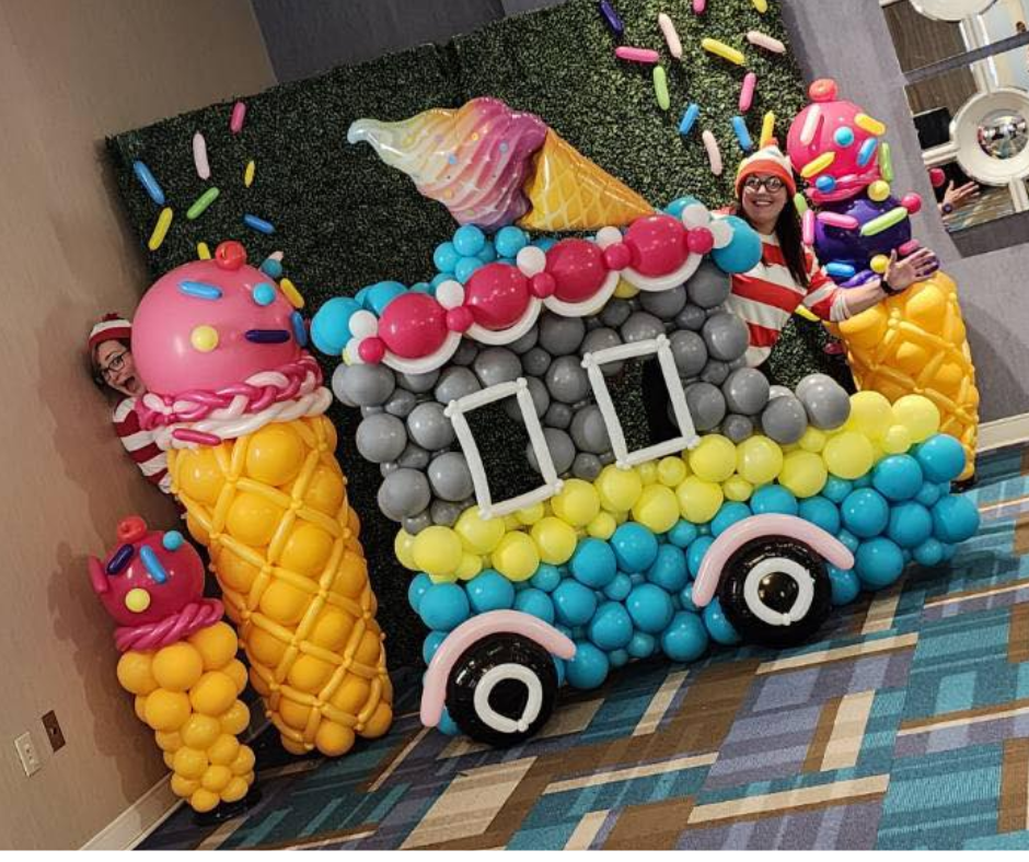 An ice cream truck made of balloons and three giant ice cream cone decorations, with someone peeking out from behind it.