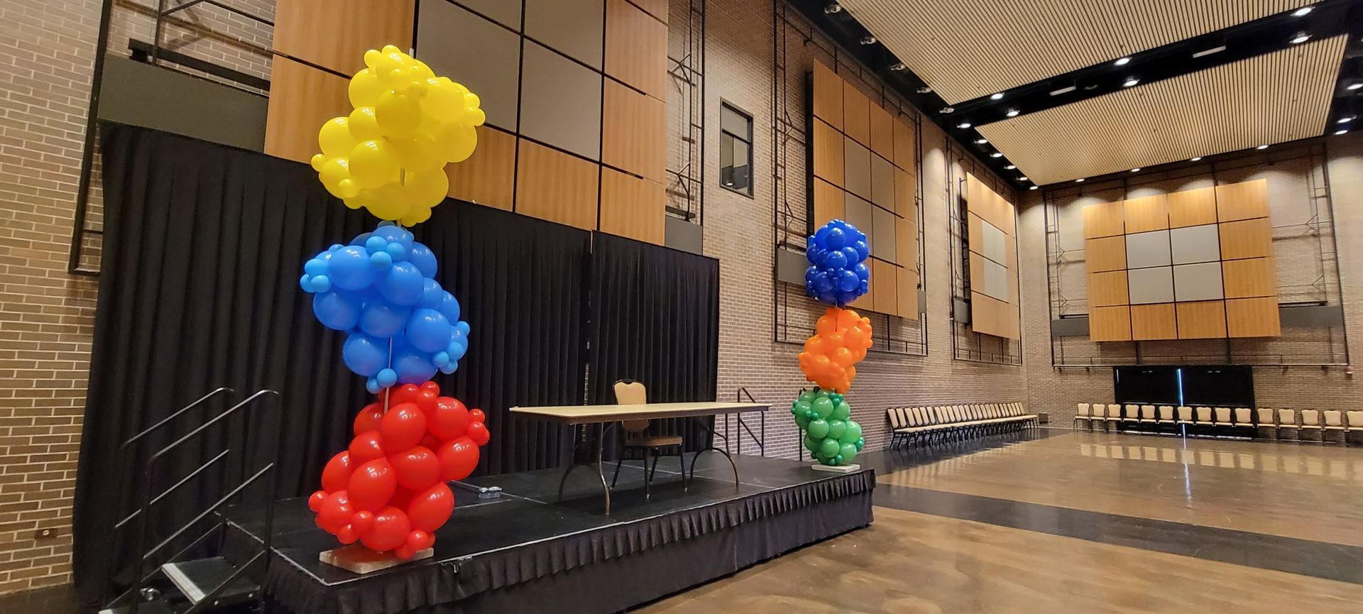 Stage decorated with balloon clusters in red, blue, yellow, orange and green in a hall.