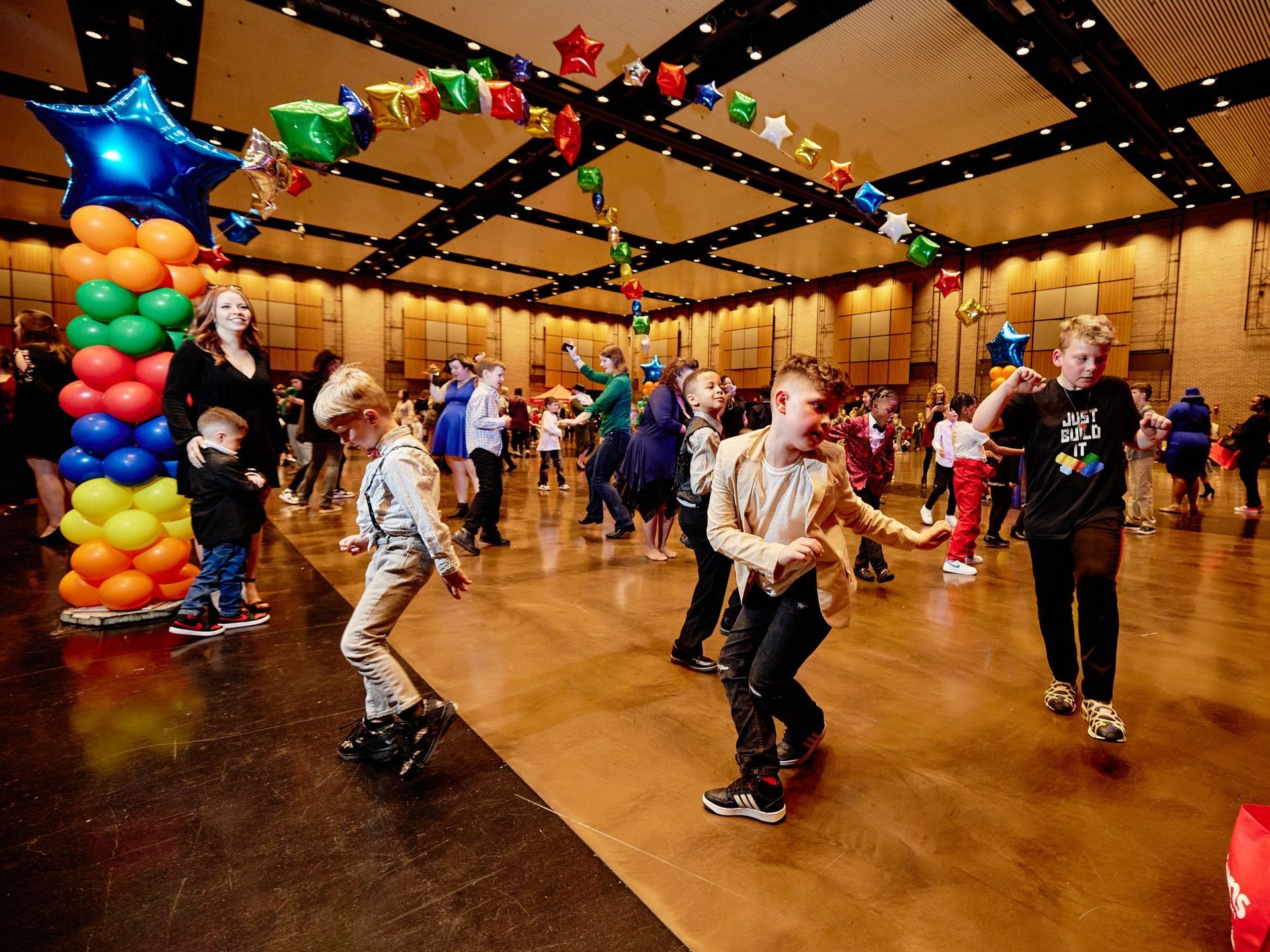 A group of children are dancing in a large room with balloons hanging from the ceiling.
