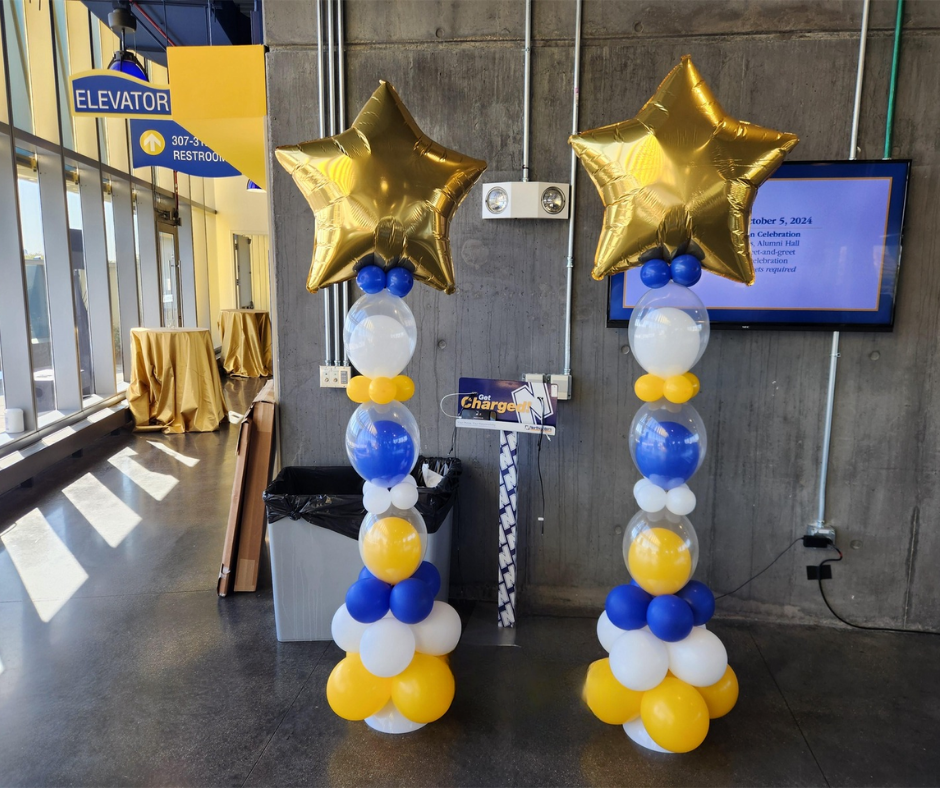 Two balloon columns with gold star toppers, blue and yellow balloons. Set in a building.