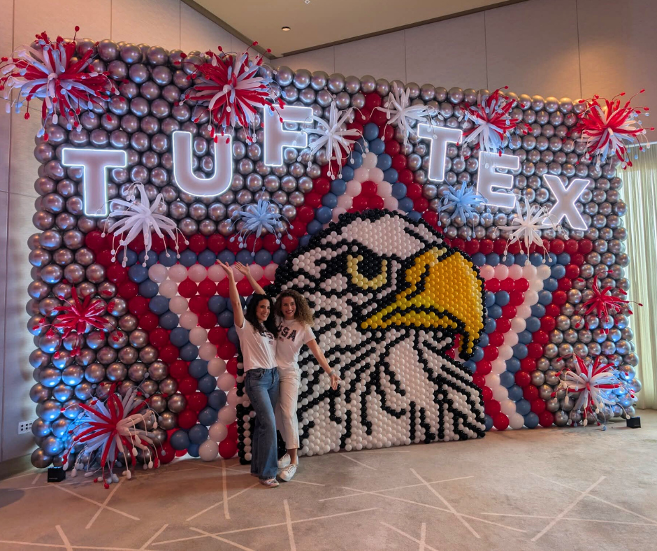 Two people pose in front of a balloon wall with a TUFTEX logo, eagle, and starburst decorations.