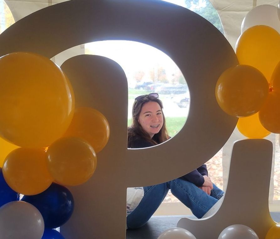 A woman sits behind a large letters for Pitt surrounded by yellow and blue balloons