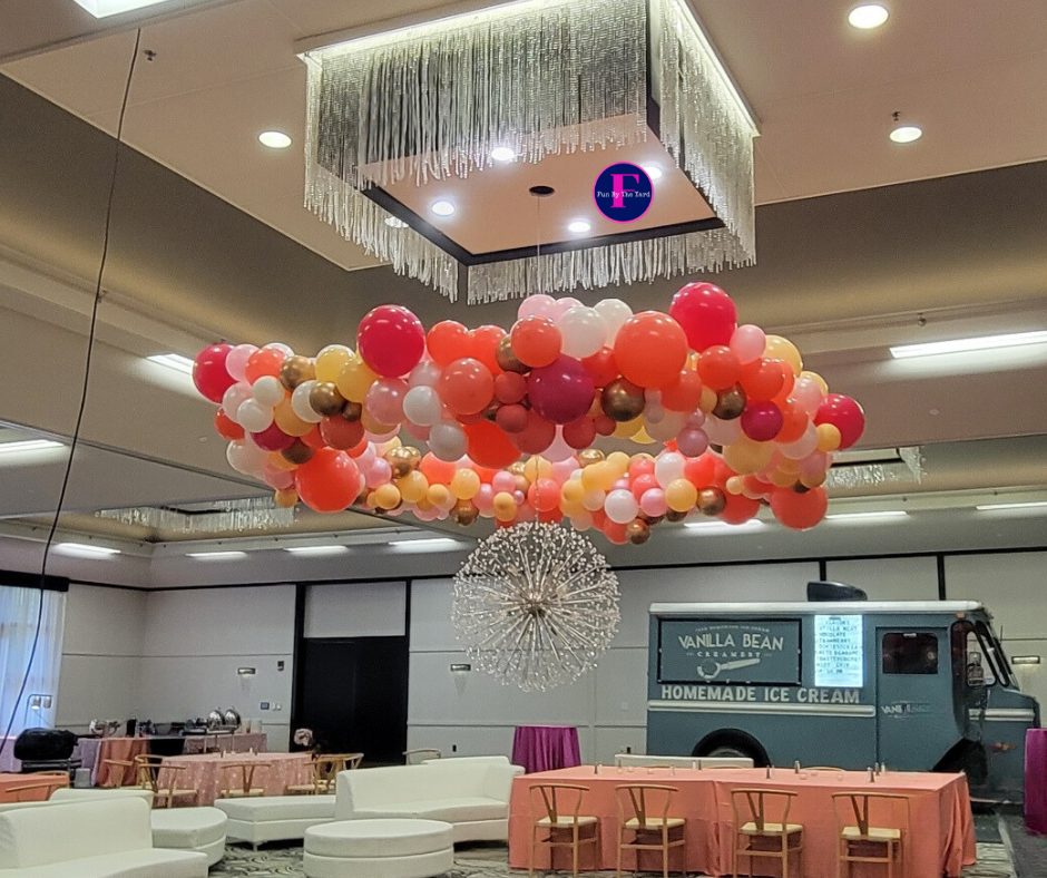 A party setting with a balloon arch in orange and white colors, with a food truck in the background.