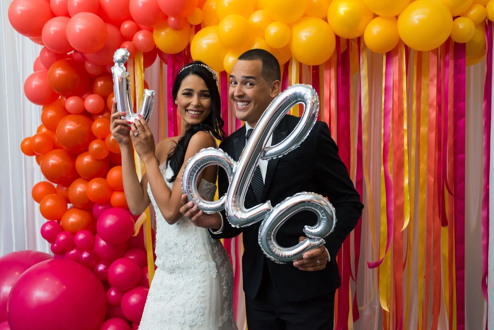 A bride and groom are holding balloons in front of a wall of balloons.