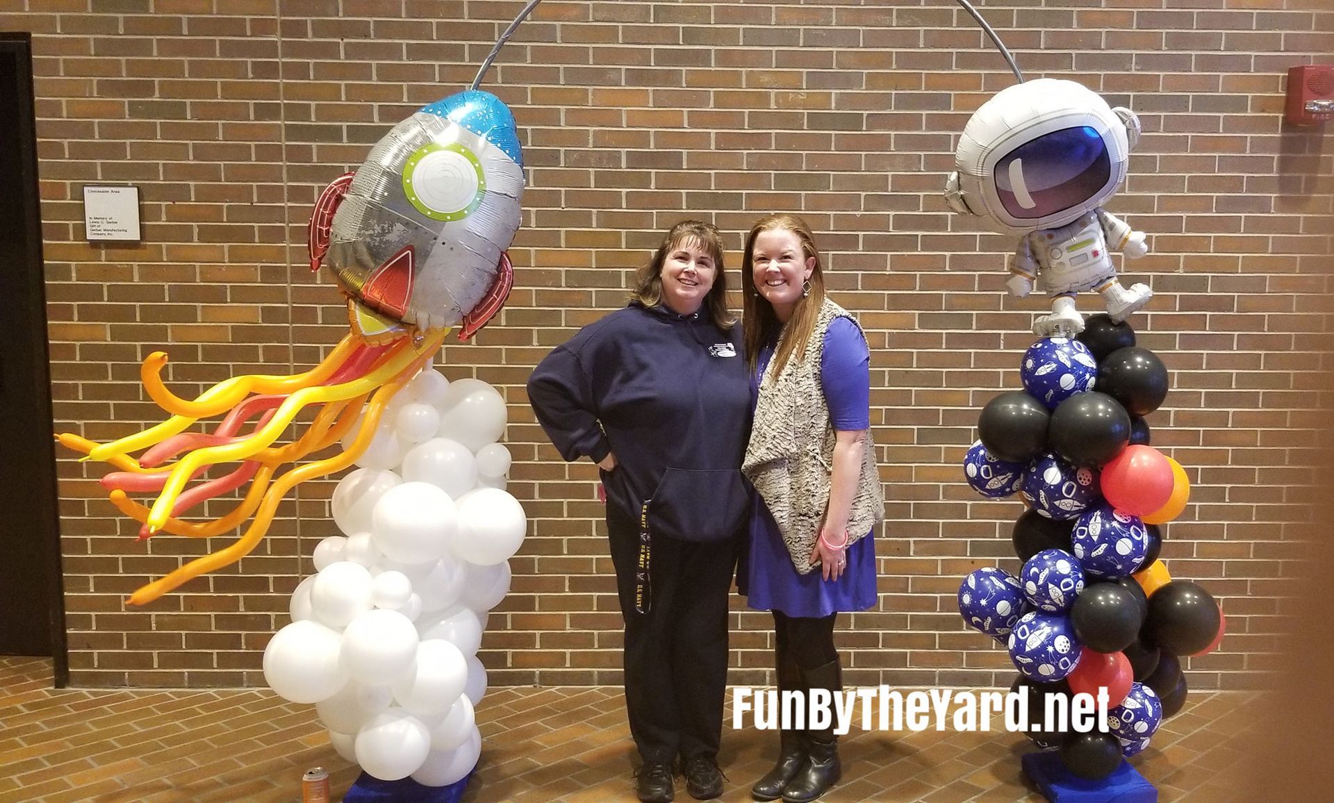 Two women are standing next to a balloon display with a rocket and an astronaut.