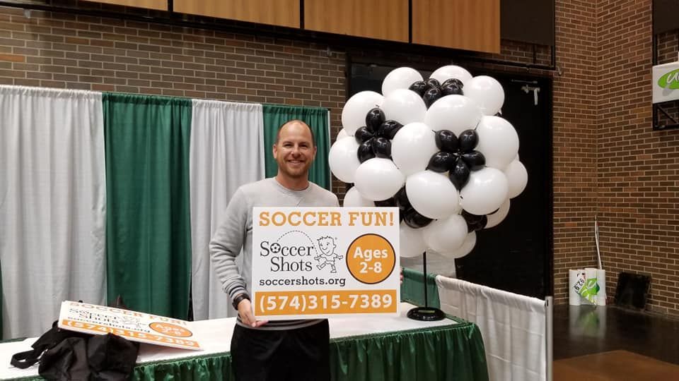 A man is standing in front of a table with balloons and a sign that says `` soccer fun ''.