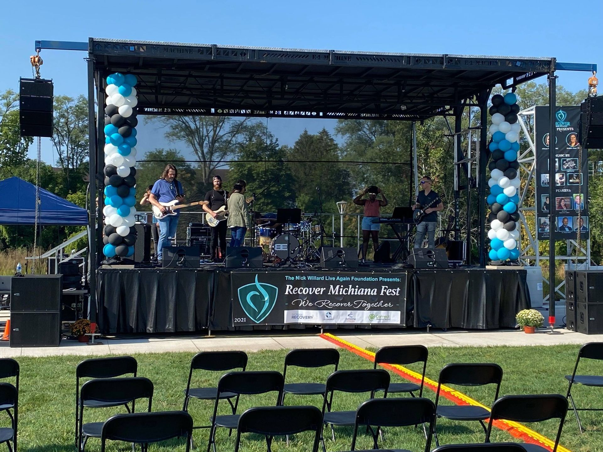 Band performing on outdoor stage; black and blue balloons, empty chairs in foreground.