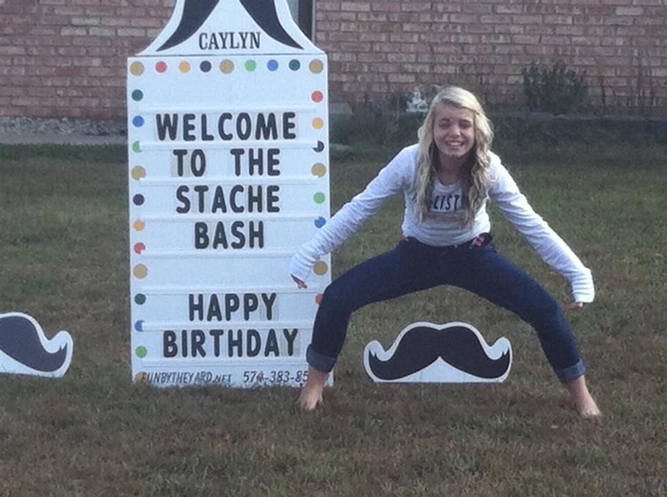 A girl is standing in front of a sign that says welcome to the stache bash happy birthday