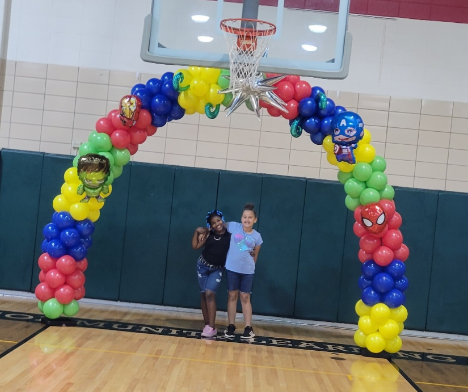 Two children are standing under a balloon arch in front of a basketball hoop.