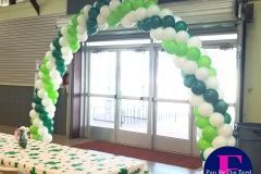 A green and white balloon arch is surrounding a table in a room.