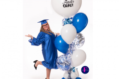 A woman in a graduation cap and gown is standing next to a bunch of blue and white balloons.
