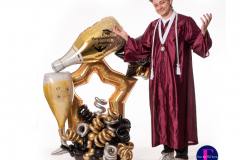 A man in a graduation gown is standing next to a balloon sculpture of a bottle of champagne.